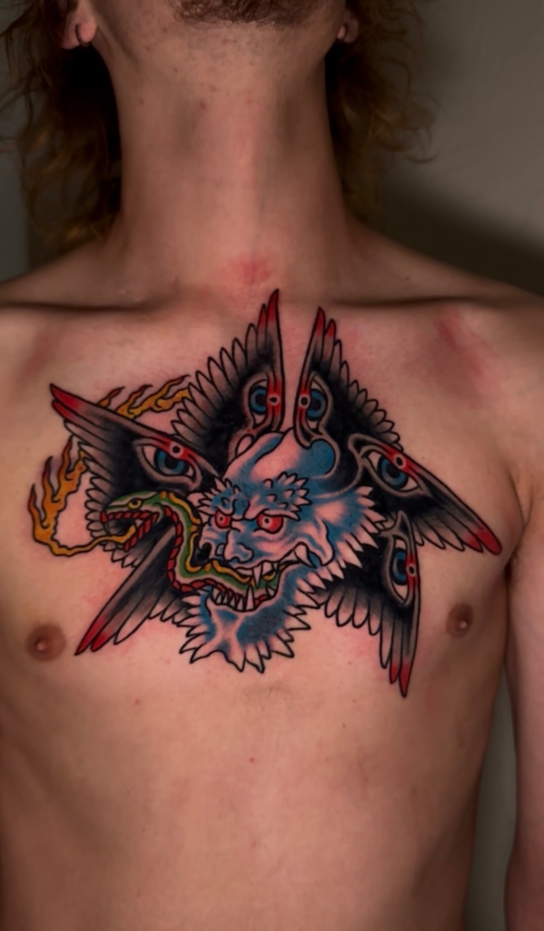 Full colour traditional tattoo of a winged horned figure holding a serpent, across the chest