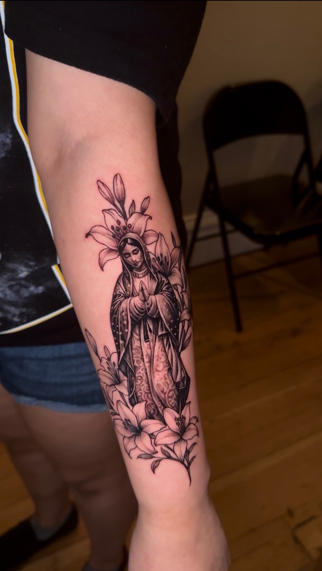 Black and grey tattoo of the Virgin Mary in prayer surrounded by lilies, on a forearm