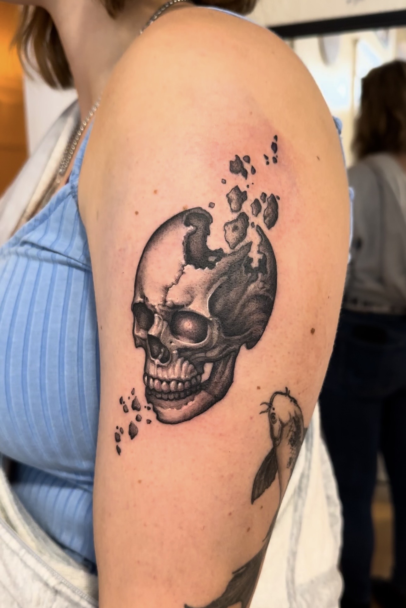 Black and grey tattoo of a skull breaking apart into floating fragments, on an upper arm